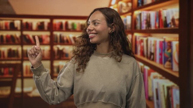 Woman points finger upward in library aisle, smiling with closed eyes and casual sweatshirt; learning curiosity joy.