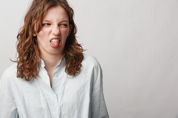 Young woman with curly hair in white shirt makes disgusted face, sticking out tongue. Isolated on...