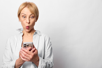 Fototapeta premium Surprised mature woman with short blond hair looks at smartphone with wide eyes and pursed lips. Isolated on white background. Concept of shock, message, and technology use.