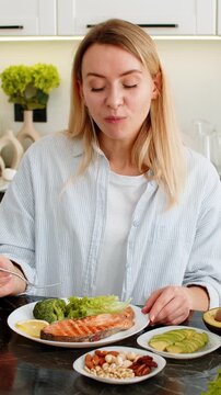 Middle-aged woman eats keto meal with fish trout avocado nuts and greens in home kitchen for healthy weight loss. Girl follows low carb nutrition plan and learns mindful eating for fitness indoors