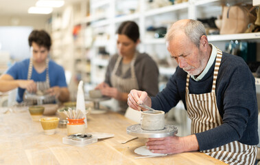 Mature man at table molds cup from wet clay in a pottery workshop