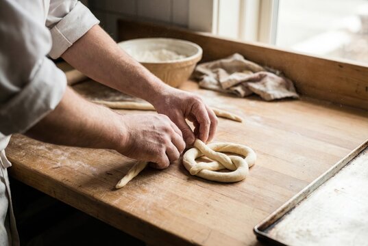 Baker hands twisting raw dough to shape a traditional pretzel on a wooden bakery table