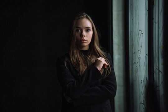 Low key portrait of a mysterious young woman in a black jacket posing near a window in the dark