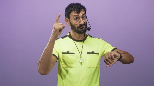 Man referee checking his watch and pointing finger, whistle on lanyard and headset mic visible, yellow referee jersey against purple studio wall; urgency.
