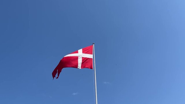 National flag of Denmark, the Dannebrog, waving proudly against bright blue sky, red banner with white Nordic cross fluttering in wind on sunny day patriotic Scandinavian emblem, horizontal footage