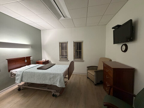 A private patient room in a skilled nursing or rehabilitation facility, shown from a wide angle. Features a hospital-style adjustable bed with a decorative headboard, overbed tray table, wall-mounted 