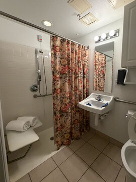 A functional accessible bathroom in a senior care or skilled nursing facility, featuring a roll-in shower with grab bars, handheld showerhead, shower chair with folded white towels, pedestal sink with