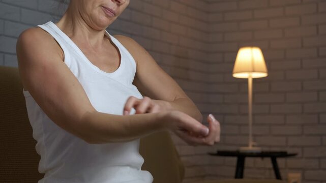 Woman scratching her itchy arm at night. Uncomfortable adult woman in her room at night, vigorously scratching an itchy arm caused by an allergic reaction, insect bite, or dermatitis, feeling pain