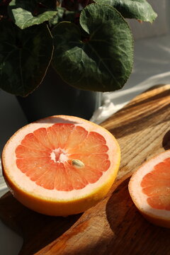 Fresh pink grapefruit halves on a rustic wooden cutting board with warm sunlight and shadows, featuring green plant leaves in the background.