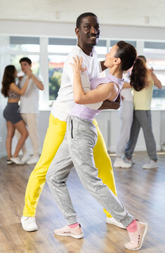 Expressive African American man in bright yellow sweatpants practicing sensual bachata with attractive graceful brunette in dance class. Social dancing concept..