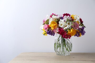Beautiful freesia flowers in glass vase on wooden table near white wall indoors, space for text