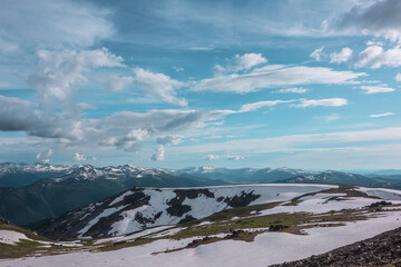 View from sunlit snowfield on hill top to snow cornice on rocky ridge against big snowbound mountain range far away under dramatic cloudy sky. Sunlight on snowy field in high mountains under clouds. © Daniil