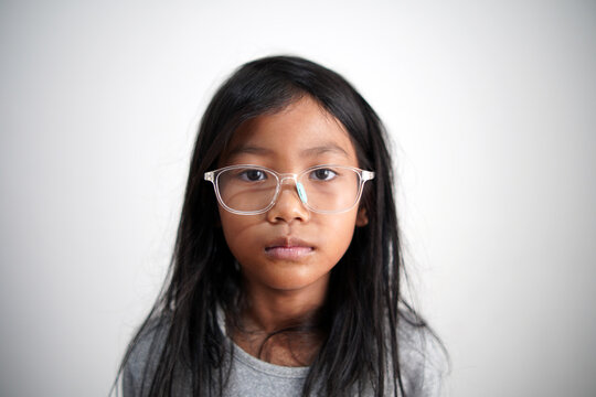 Little Asian girl with eyeglasses looking at camera with neutral expression, standing indoors, blank stare, serious childhood face