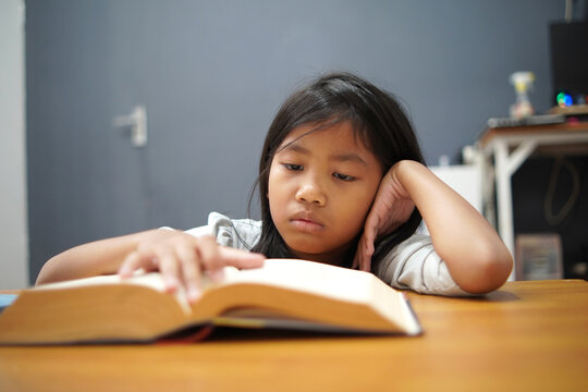 Bored little Asian girl leaning on hand while reading a thick book, tired child feeling unmotivated or sleepy during homework, academic boredom and learning difficulties