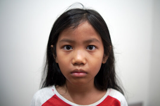 Little Asian girl looking at camera with neutral expression, standing indoors, blank stare, serious childhood face