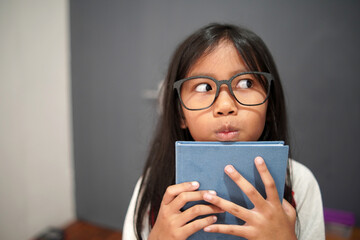 Fototapeta premium Thoughtful little Asian girl with glasses holding a book and looking away, curious child student thinking of an idea, imagination, creativity, and education