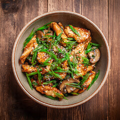 Fototapeta premium Stir fry chicken with paprika, mushrooms, green chives and sesame seeds in ceramic bowl. Asian cuisine dish. Wooden kitchen table background, top view