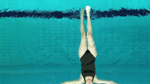 Top down view of a female athlete in a black swimsuit pushing off the wall to perform a backstroke start in a clear blue pool