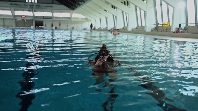 Female scuba diver in full gear surfacing in an indoor swimming pool, holding an underwater camera after a dive training session