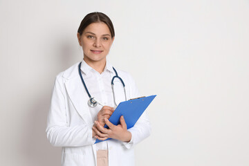 Fototapeta premium Portrait of smiling doctor in medical uniform with stethoscope and clipboard on light background. Space for text