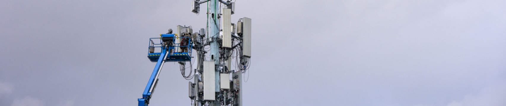 Boom truck lift backet with technician workmen raised up to antenna platform to perform maintenance, wireless mobile communication co-located cell sites on tall tower on cold winter day
