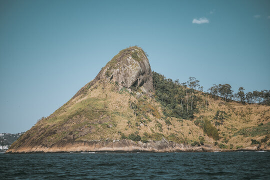 Telephoto view of rocky coastal island hill rising from dark water, with scattered trees and rugged cliff face, clear blu e sky, natural shoreline landscape; Parque Dora Negreiros, Parque do Morcego