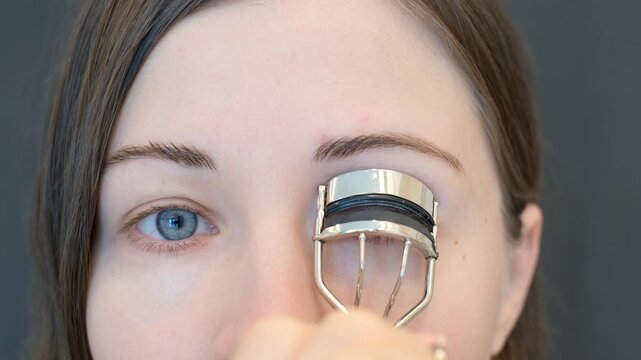 Young woman using eyelash curler on grey background close-up.