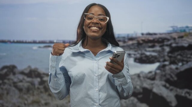 Woman holding smartphone and pumping fist while smiling in a studio with rocky seaside backdrop and eyeglasses visible; celebration success.