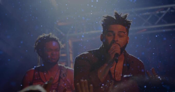 Singer starting set on music stage leaning, emoting for crowd, bandmate playing, particles drifting