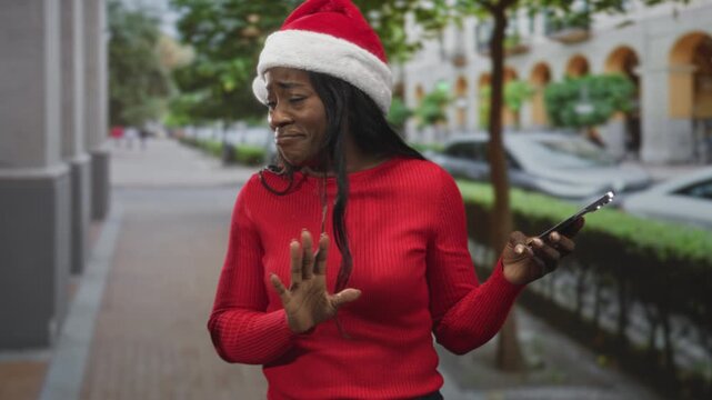 Woman in santa hat holds smartphone, raises hand in a defensive grimace and recoils on street near buildings and parked cars; disgust surprise.