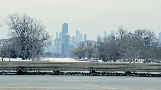 Tall buildings of Chicago stand in the background behind an icy lake and bare trees, views from a road in winter. The roads are clear of snow, showing signs of urban life.