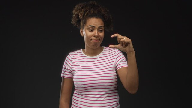 Woman pinches fingers close in black studio while wearing a white and pink striped tee and showing a wry expression; skepticism.