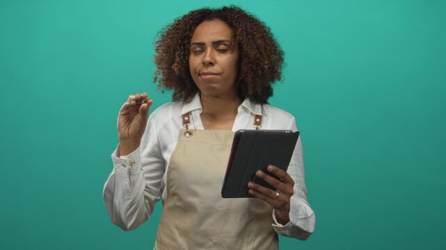 Young african american woman baker in beige apron holding tablet, left hand pinching gesture while checking recipe in teal studio; focus skill.