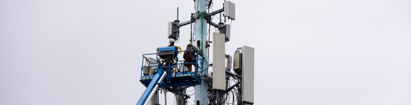 Boom truck lift backet with technician workmen raised up to antenna platform to perform maintenance, wireless mobile communication co-located cell sites on tall tower on cold winter day
