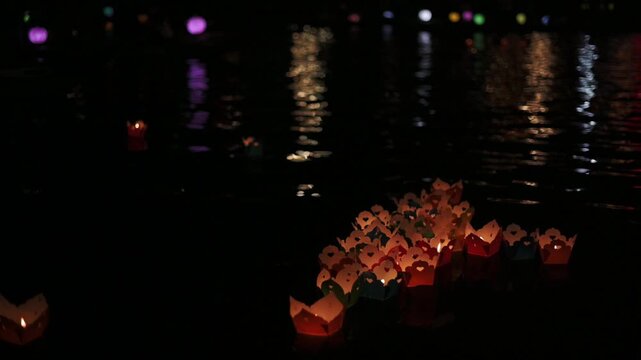 Bright Processional Display With Floating Candles Under City Reflections At Night. Colorful Lantern Procession Gently Moves Across Rippled Water Reflecting Vibrant City Lights At Dusk