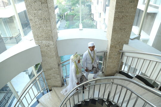 Indonesian bride and groom in traditional white wedding attire standing together on a modern staircase, sharing a quiet romantic moment before the ceremony.