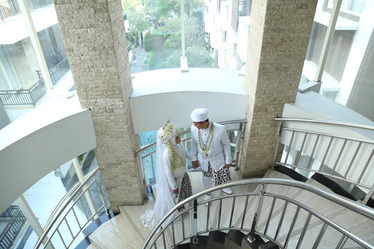 Indonesian bride and groom in traditional white wedding attire standing together on a modern staircase, sharing a quiet romantic moment before the ceremony.