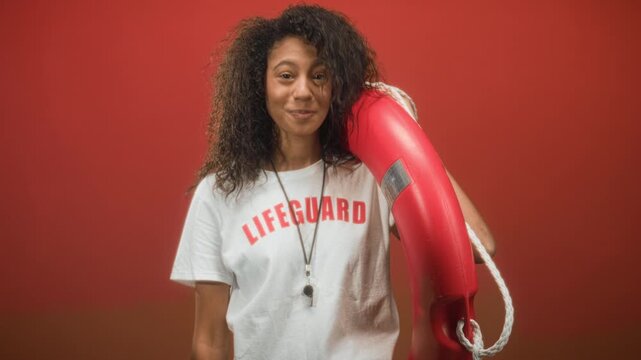 Young woman lifeguard holding red lifebuoy on her shoulder and smiling in studio; confidence readiness.