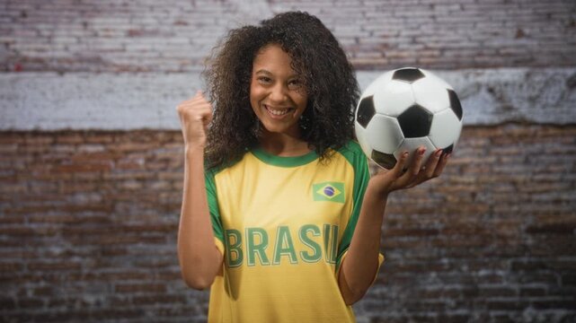Teenage woman holding soccer ball and pumping fist while smiling, wearing brasil jersey with flag patch in studio; pride celebration support.