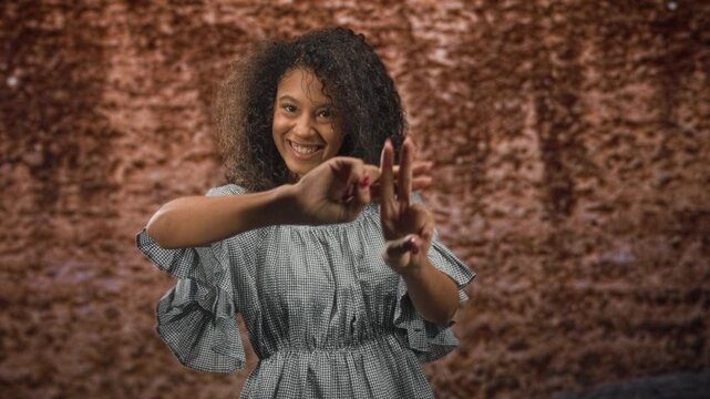 Woman forming a hashtag sign with her hands while smiling, wearing a gingham dress and curly hair against a textured backdrop in a studio setting; joy self expression.