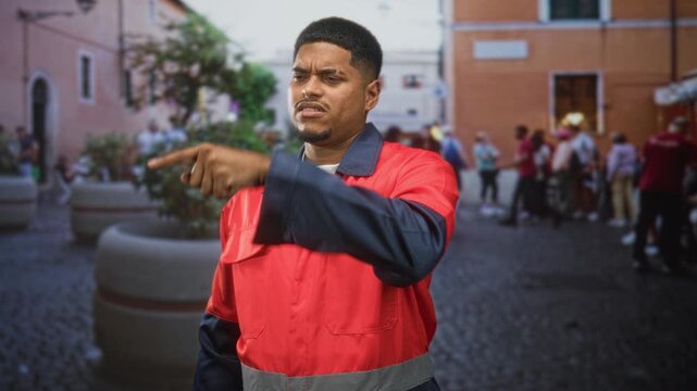 Man with beard in red worker uniform pointing finger toward a crowd on cobblestone street plaza near planters and building facade; authority.