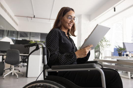 Positive young Middle Eastern female project manager in wheelchair using professional online service on tablet, typing on screen, smiling, working for company with accessibility