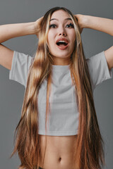Fototapeta premium Young woman with long hair and braids, surprised expression, wearing white crop top, posing with hands on head against gray background indoors.