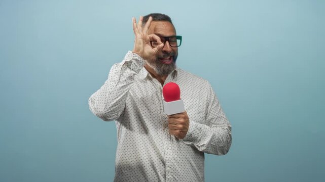 Man reporter, middle aged hispanic with grey beard and glasses, holding red microphone and making ok hand gesture in light blue studio; confidence.