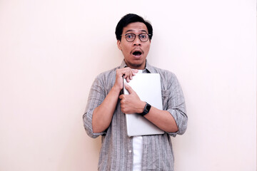 Man shocked, holding laptop, against plain white wall. Eyes wide, mouth agape, conveying surprise...