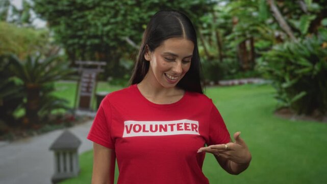 Young brunette woman in a red volunteer t shirt points to the logo and smiles while standing in a forest path with green foliage; joyful service.