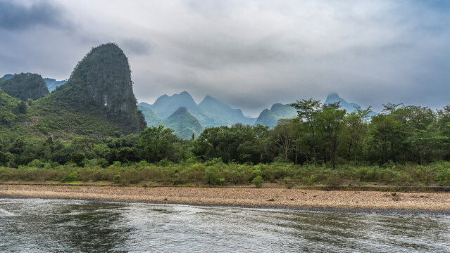 A picturesque calm river. There is green vegetation on the pebbly shore. A mountain range on a background of sky and clouds. China. Li River. Li Jiang. Guilin