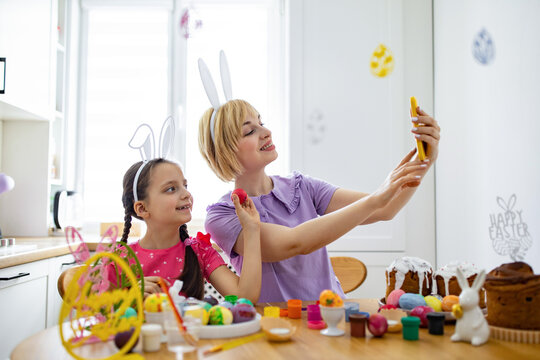 A mother and daughter wearing bunny ears take a selfie with painted Easter eggs and cakes on the table