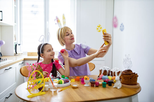 A mother and daughter wearing bunny ears take a selfie while decorating Easter eggs and cakes in a bright kitchen