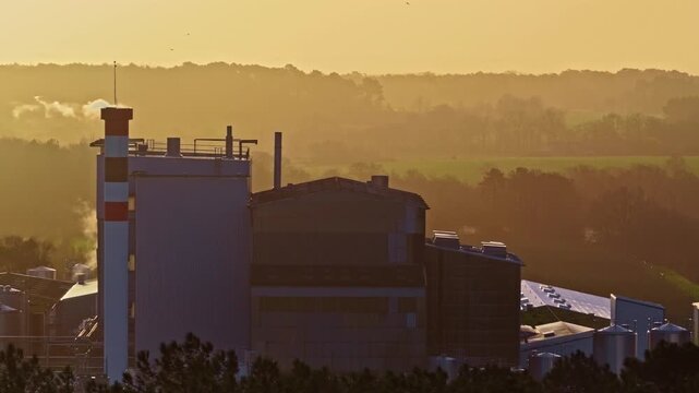 Golden sunset glow over large industrial factory complex with tall chimneys emitting steam silos and buildings surrounded by fields and forest in Jelgava Latvia